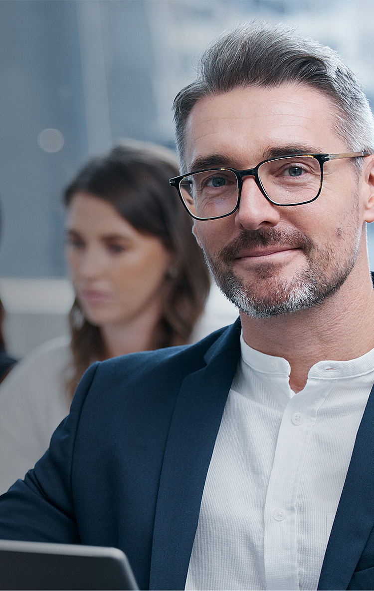 Ein lächelnder Mann mit Brille in einem modernen Büro, gekleidet in einem Anzug. Er sitzt am Tisch, während zwei Frauen im Hintergrund an Laptops arbeiten. Die Atmosphäre ist professionell und konzentriert.