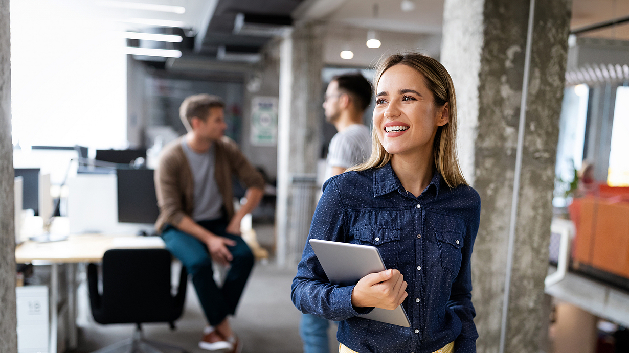 Eine junge Frau lächelt, während sie in einem modernen Büro steht und ein Laptop in der Hand hält. Im Hintergrund sind mehrere Büros und andere Personen zu sehen. Die Frau trägt ein blaues Hemd und gelbe Hosen, was ihr ein frisches Aussehen verleiht.